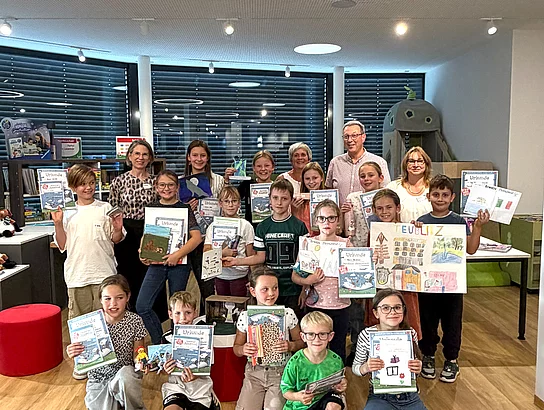 Eine Gruppe von Kindern steht und kniet gemeinsam mit dem Büchereiteam und Stadtkämmerer Georg Beer in der Stadtbücherei und präsentiert Urkunden sowie selbst gestaltete Bilder und Arbeiten.