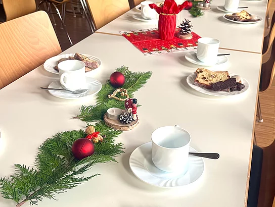 Lange Tische in einem Saal sind für eine Weihnachtsfeier gedeckt mit Kaffeetassen, Tellern mit Gebäck und weihnachtlicher Dekoration aus Tannenzweigen, roten Kugeln und kleinen Figuren.