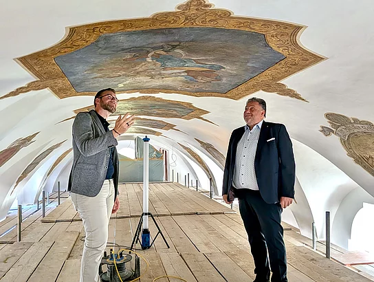 Pfarrer Lucas Lobmeier und Bürgermeister Thomas Beer stehen auf dem großflächigen Arbeitsgerüst im Gewölbe der Marien-Wallfahrtskirche Saltendorf. Lobmeier blickt nach oben und deutet mit einer Hand auf die historischen Deckengemälde, während Beer ihm gegenübersteht. Mehrere der gerahmten Gemälde sind über den beiden zu sehen.