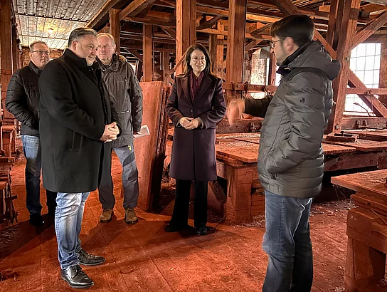 Eine kleine Gruppe von Personen lässt sich in einem historischen, hölzernen Werkstattgebäude etwas erklären und steht aufmerksam zwischen alten Maschinen und Balkenkonstruktionen.