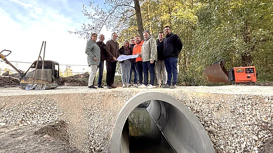 Acht Personen stehen bei einer Baustellenbesichtigung am Grünwinkelgraben auf einem neu angelegten Wirtschaftsweg. Einige von ihnen halten gemeinsam einen Bauplan in den Händen. Im Vordergrund ist der große, neu eingebaute Betondurchlass zu sehen, durch den der Bach fließt. Rund um das Bauwerk liegen Schotter und Erdreich. Im Hintergrund sind Bäume und Baumaschinen zu erkennen.