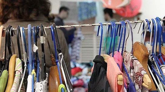 Verschiedene bunte Klamotten aufgehängt auf Bügeln und an einer Stange hängend. im Hintergrund ein bunter Verkaufsraum.