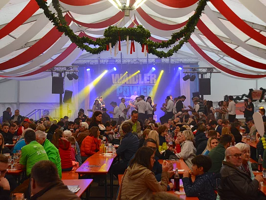 Blick in ein gut gefülltes Bierzelt mit rot-weißem Himmel, Kronleuchter und einer vorbereiteter Bühne.