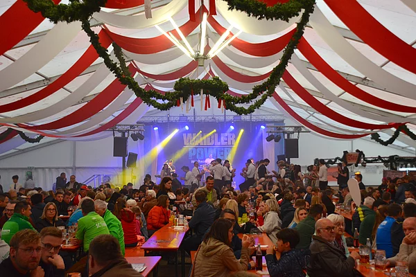 Blick in ein gut gefülltes Bierzelt mit rot-weißem Himmel, Kronleuchter und einer vorbereiteter Bühne.