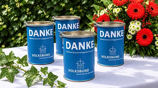Vier blaue Sammeldosen des Volksbundes Deutsche Kriegsgräberfürsorge stehen auf einem weißen Tisch. Auf den Dosen sind in weißer Schrift „DANKE“ sowie das Volksbund-Logo und der Schriftzug „Gemeinsam für den Frieden“ zu sehen. Im Hintergrund befinden sich grüner Efeu und ein Blumenarrangement aus roten und weißen Blüten.