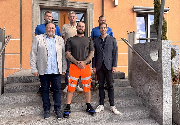 Sechs Männer stehen für ein Gruppenfoto auf der Treppe vor einem orangefarbenen Gebäude. Im Mittelpunkt steht der neue Bauhofmitarbeiter Patrick Mailbeck in auffälliger orangefarbener Arbeitshose. Neben und hinter ihm stehen fünf Männer, die ihn zum Beginn seiner neuen Tätigkeit willkommen heißen.
