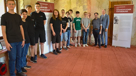 Eine Gruppe von Schülerinnen und Schülern sowie zwei Lehrkräfte stehen in den historischen Räumen der Münchshofener „Schleif“ zwischen zwei Rollups mit Informationen zur Geschichte des ehemaligen Glasschleif- und Polierwerks und präsentieren die Ergebnisse ihres P-Seminars.
