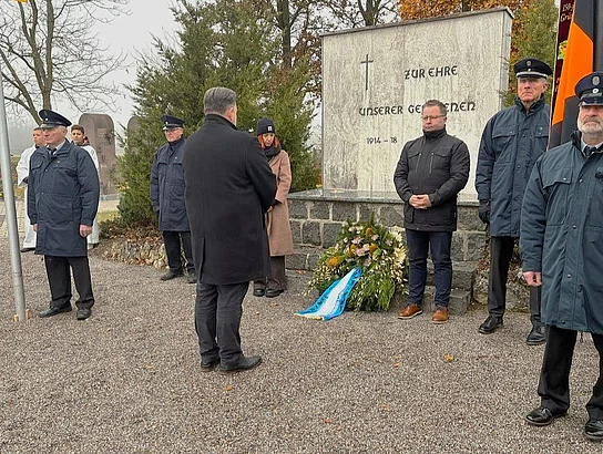 Mehrere Menschen stehen in Uniform und Zivil vor einem Kriegerdenkmal, während ein Kranz mit blau-weißem Band davor niedergelegt ist.