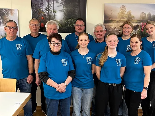 Eine Gruppe von Frauen und Männern in einheitlichen blauen T-Shirts steht nebeneinander und blickt lächelnd in die Kamera, offenbar als Teamfoto eines Service- oder Helferteams.