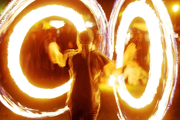 Eine Künstlerin präsentiert bei Dunkelheit eine spektakuläre Feuershow. Durch kreisende Feuergeräte entstehen zwei große leuchtende Feuerringe, die den Körper der Artistin eindrucksvoll umrahmen. Im Hintergrund sind weitere Flammen und schemenhaft Zuschauer zu erkennen.