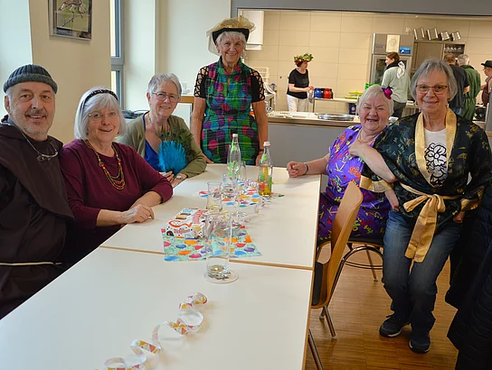 Eine fröhliche Gruppe älterer Menschen, die Mitglieder des Seniorenbeirats sind, sitzt an einem Tisch, lächelt in die Kamera und trägt teilweise farbenfrohe Kleidung und Faschingsaccessoires.