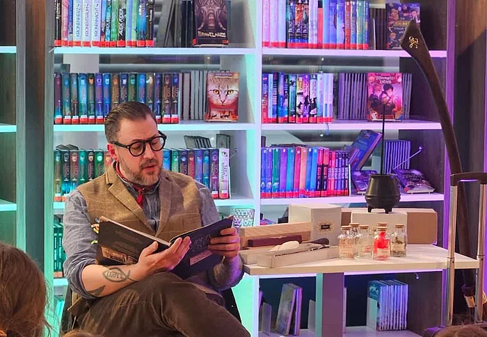 Ein Vorleser sitzt mit einem geöffneten Buch vor einer Gruppe von Kindern und liest ausdrucksstark vor. Neben ihm stehen Requisiten wie kleine Fläschchen und ein schwarzer Kessel auf einem Tisch. Die farbig beleuchteten Bücherregale im Hintergrund sorgen für eine besondere Atmosphäre.