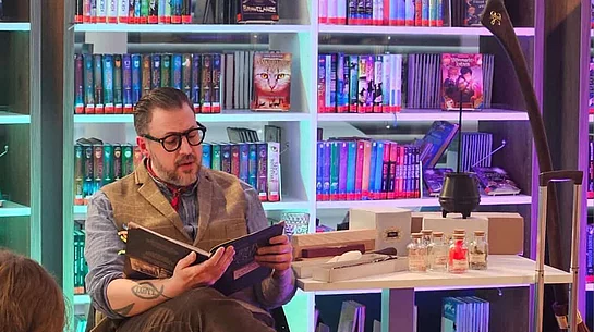 Ein Vorleser sitzt mit einem geöffneten Buch vor einer Gruppe von Kindern und liest ausdrucksstark vor. Neben ihm stehen Requisiten wie kleine Fläschchen und ein schwarzer Kessel auf einem Tisch. Die farbig beleuchteten Bücherregale im Hintergrund sorgen für eine besondere Atmosphäre.