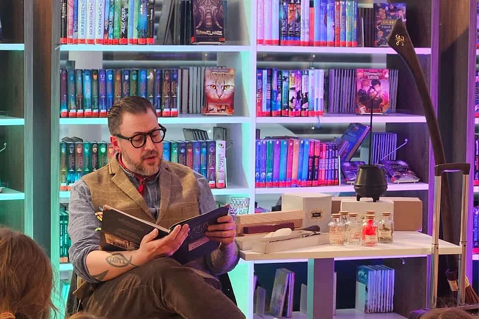 Ein Vorleser sitzt mit einem geöffneten Buch vor einer Gruppe von Kindern und liest ausdrucksstark vor. Neben ihm stehen Requisiten wie kleine Fläschchen und ein schwarzer Kessel auf einem Tisch. Die farbig beleuchteten Bücherregale im Hintergrund sorgen für eine besondere Atmosphäre.