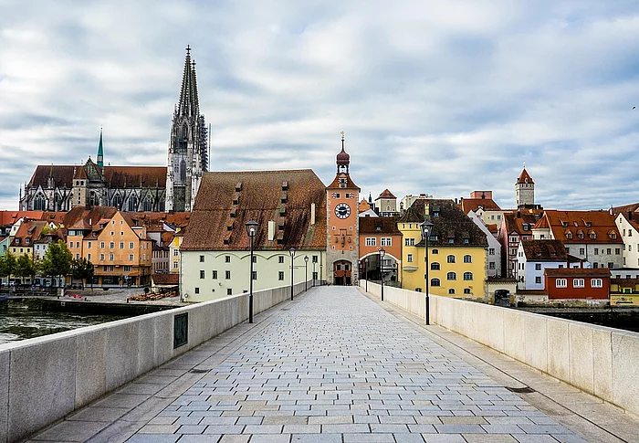 Eine gepflasterte Brücke führt auf eine historische Altstadt mit bunten Häusern, einem Stadttor und dem Dom im Hintergrund unter bewölktem Himmel zu.