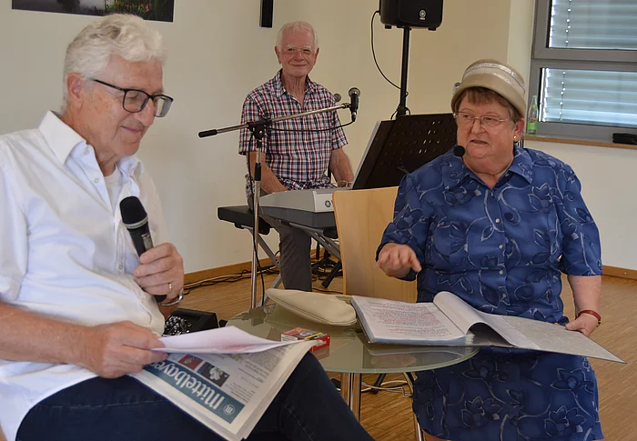 In einem Veranstaltungsraum sitzen ein Mann mit Mikrofon und Unterlagen sowie eine Frau mit geöffnetem Ordner an einem kleinen Tisch, während im Hintergrund ein weiterer Mann an einem Keyboard sitzt und die Szene musikalisch begleitet.
