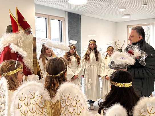 Mehrere Kinder in weißen Engelskostümen mit goldenen Heiligenscheinen und Flügeln stehen in einem Raum zusammen mit einem als Nikolaus verkleideten Erwachsenen. Ein Mann auf der rechten Seite nimmt ein Geschenkset entgegen und lächelt. Die Szene wirkt wie ein festlicher Besuch oder eine Verabschiedung im Rahmen einer Nikolausaktion.
