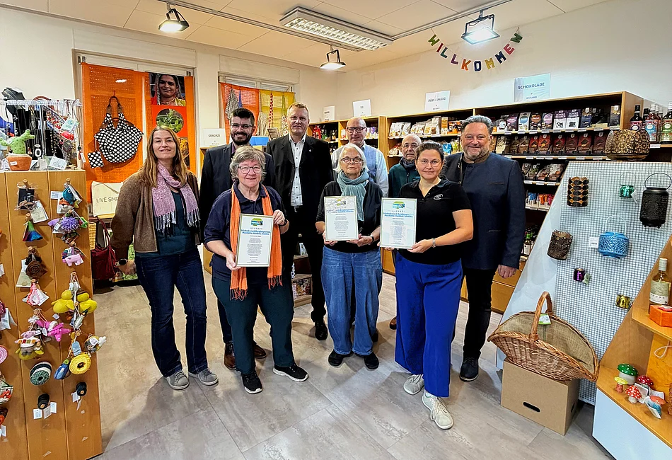 Gruppenfoto im Weltladen Teublitz anlässlich der Verlängerung der Fairtrade-Auszeichnung für das Städtedreieck bis 2028. Neun Personen, fünf Männer und vier Frauen, stehen in zwei Reihen vor Verkaufsregalen mit fair gehandelten Lebensmitteln und Produkten. Drei Frauen in der vorderen Reihe halten gerahmte Urkunden zur erneuten Auszeichnung in den Händen. Im Hintergrund sind Regale mit Schokolade, Tee und weiteren Waren sowie eine bunte Willkommensgirlande zu sehen. Die Gruppe blickt in die Kamera.