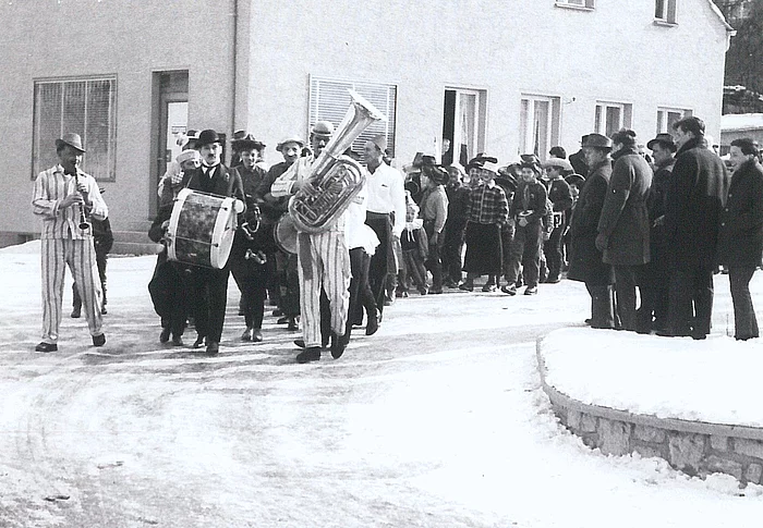 Das historische Schwarzweißfoto zeigt einen Faschingszug im Jahr 1958, bei dem eine Musikgruppe in gestreiften Kostümen voran marschiert, gefolgt von zahlreichen Zuschauern und Teilnehmern entlang einer verschneiten Straße.