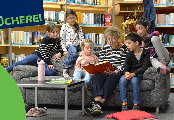 Ein Werbebild für die Stadtbücherei zeigt eine ältere Frau, die mehreren Kindern auf einem Sofa aus einem Buch vorliest. Die Kinder sitzen aufmerksam um sie herum und hören gespannt zu. Im Hintergrund sind Bücherregale der Bücherei zu sehen. Links oben steht gut sichtbar der Schriftzug „BÜCHEREI“.