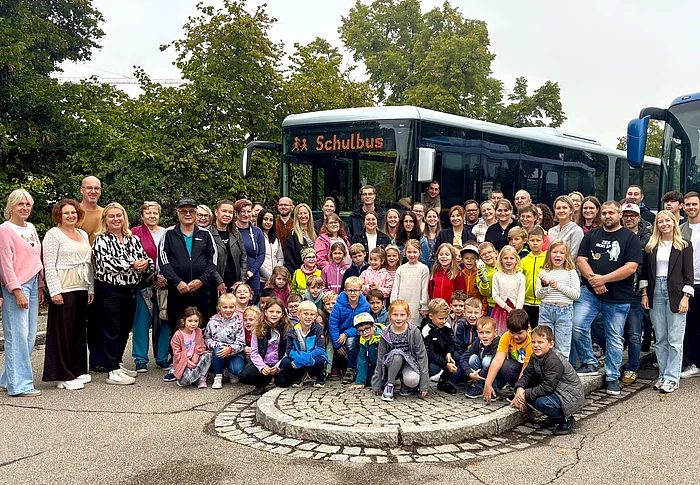 Auf dem Foto ist eine große Gruppe von Menschen zu sehen – Erwachsene und Kinder – die gemeinsam vor zwei Bussen stehen. Auf einem der Busse steht leuchtend „Schulbus“.