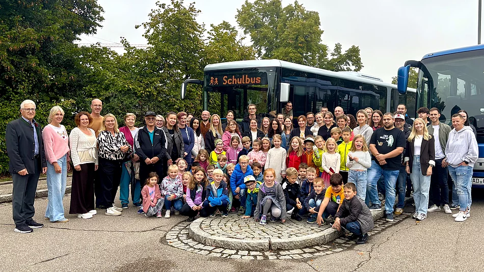 Auf dem Foto ist eine große Gruppe von Menschen zu sehen – Erwachsene und Kinder – die gemeinsam vor zwei Bussen stehen. Auf einem der Busse steht leuchtend „Schulbus“.