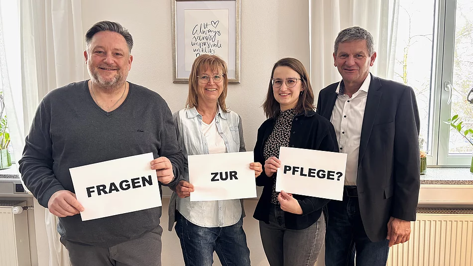 Vier Personen stehen nebeneinander in einem hellen Raum und halten jeweils ein Blatt Papier hoch, sodass zusammen die Frage „Fragen zur Pflege?“ entsteht, während sie freundlich in die Kamera blicken.