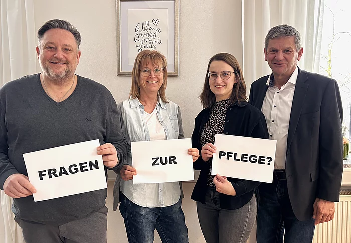 Vier Personen stehen nebeneinander in einem hellen Raum und halten jeweils ein Blatt Papier hoch, sodass zusammen die Frage „Fragen zur Pflege?“ entsteht, während sie freundlich in die Kamera blicken.