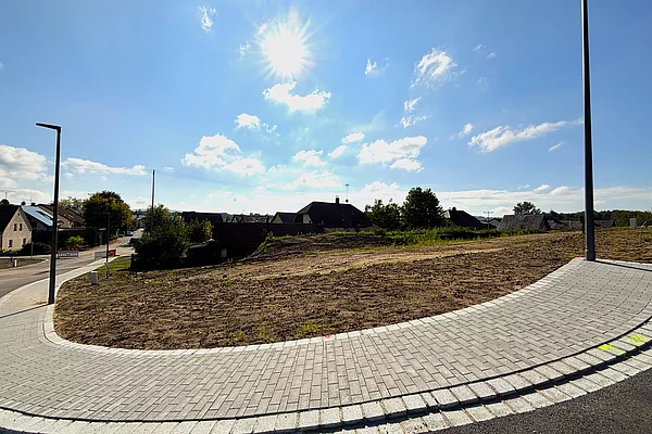 Ein leicht ansteigendes, unbebautes Grundstück mit brauner Erde liegt an einer neu asphaltierten Straße; im Hintergrund stehen einzelne Häuser am Waldrand unter blauem Himmel.