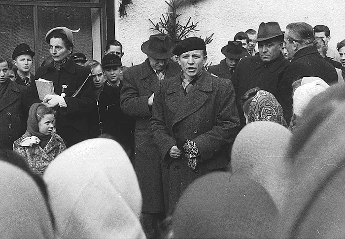 Eine historische Schwarz-Weiß-Aufnahme zeigt einen Redner mit Baskenmütze, der während der Eröffnungsfeier vor einer großen Menschenmenge spricht. Um ihn herum stehen Erwachsene und Jugendliche, während zahlreiche Besucherinnen und Besucher aufmerksam zuhören.