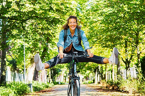 Eine lachende Frau fährt auf einem Fahrrad durch eine von Bäumen gesäumte Allee. Während der Fahrt streckt sie beide Beine seitlich aus und balanciert freihändig auf dem Fahrrad. Sie trägt eine Jeansjacke, Turnschuhe und einen kleinen Rucksack. Die Aufnahme vermittelt Freude an Bewegung und Radfahren in der Natur.