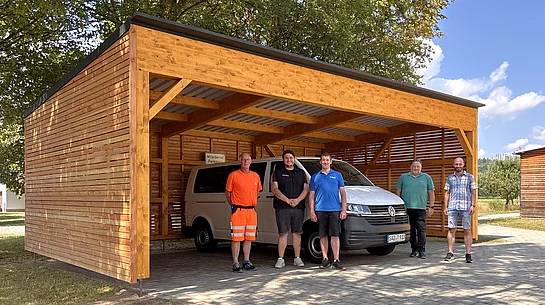 Fünf Mitarbeiter der Stadt Teublitz stehen vor dem neu errichteten Holz-Carport am Mehrgenerationenhaus, unter dem das Bürgermobil geschützt abgestellt ist. Das moderne Carport mit Holzverkleidung bietet Platz für das Fahrzeug und fügt sich harmonisch in die parkähnliche Umgebung ein.