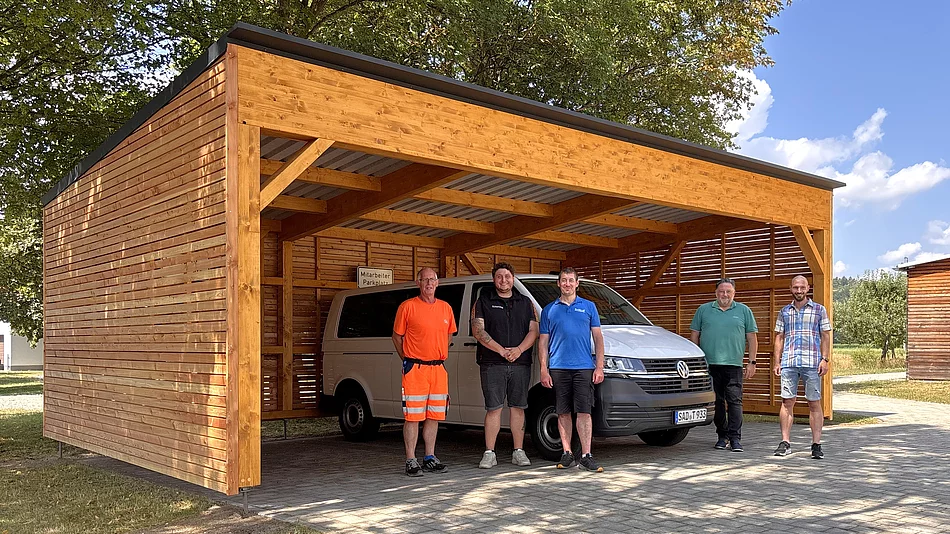 Fünf Mitarbeiter der Stadt Teublitz stehen vor dem neu errichteten Holz-Carport am Mehrgenerationenhaus, unter dem das Bürgermobil geschützt abgestellt ist. Das moderne Carport mit Holzverkleidung bietet Platz für das Fahrzeug und fügt sich harmonisch in die parkähnliche Umgebung ein.