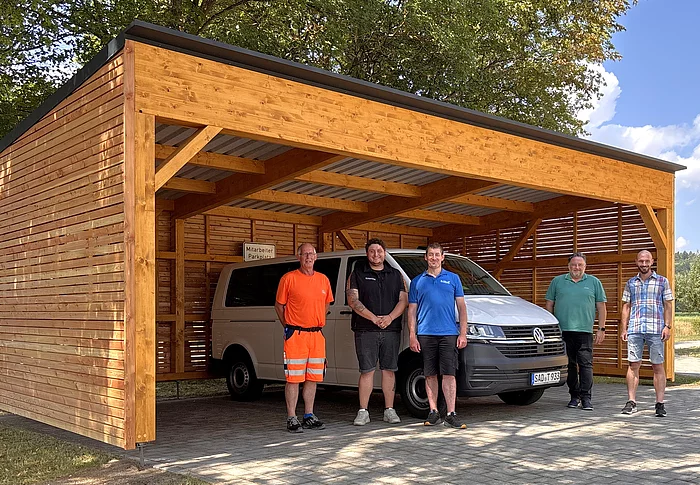 Fünf Mitarbeiter der Stadt Teublitz stehen vor dem neu errichteten Holz-Carport am Mehrgenerationenhaus, unter dem das Bürgermobil geschützt abgestellt ist. Das moderne Carport mit Holzverkleidung bietet Platz für das Fahrzeug und fügt sich harmonisch in die parkähnliche Umgebung ein.