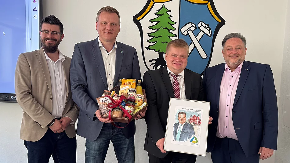 Vier Männer stehen vor einem Wappen, überreichen Geschenke und ein gerahmtes Bild und posieren gemeinsam für ein Foto.