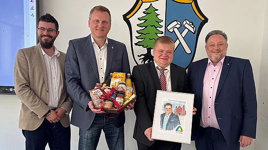 Vier Männer stehen vor einem Wappen, überreichen Geschenke und ein gerahmtes Bild und posieren gemeinsam für ein Foto.