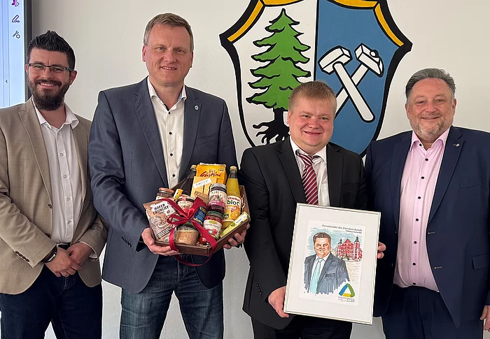 Vier Männer stehen vor einem Wappen, überreichen Geschenke und ein gerahmtes Bild und posieren gemeinsam für ein Foto.