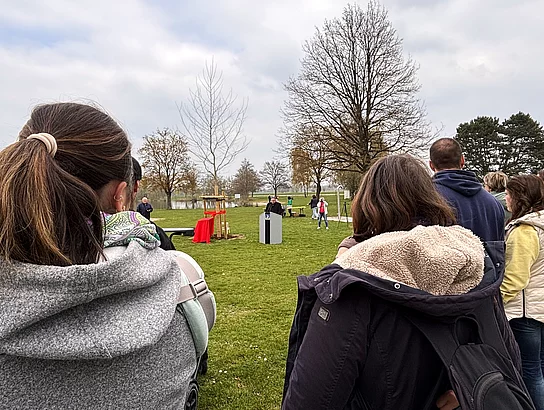 Besucherinnen und Besucher stehen auf einer Wiese und hören einer Ansprache an einem Rednerpult bei einer Baumpflanzaktion zu.