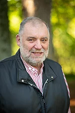 Ein älterer Mann mit grauem Bart und Weste blickt mit leichtem Lächeln in die Kamera.