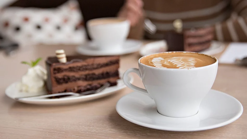 Auf einem Tisch stehen mehrere weiße Kaffeetassen und Teller mit Kuchen. Im Vordergrund ist eine Tasse Kaffee mit Milchschaum zu sehen, daneben ein Stück Schokoladenkuchen mit Sahne. Das Symbolbild steht für den gemeinsamen Seniorennachmittag des Landkreises Schwandorf.