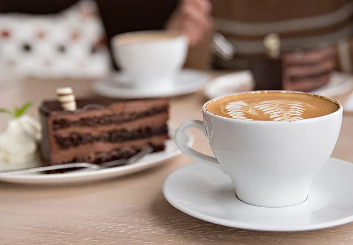 Auf einem Tisch stehen mehrere weiße Kaffeetassen und Teller mit Kuchen. Im Vordergrund ist eine Tasse Kaffee mit Milchschaum zu sehen, daneben ein Stück Schokoladenkuchen mit Sahne. Das Symbolbild steht für den gemeinsamen Seniorennachmittag des Landkreises Schwandorf.