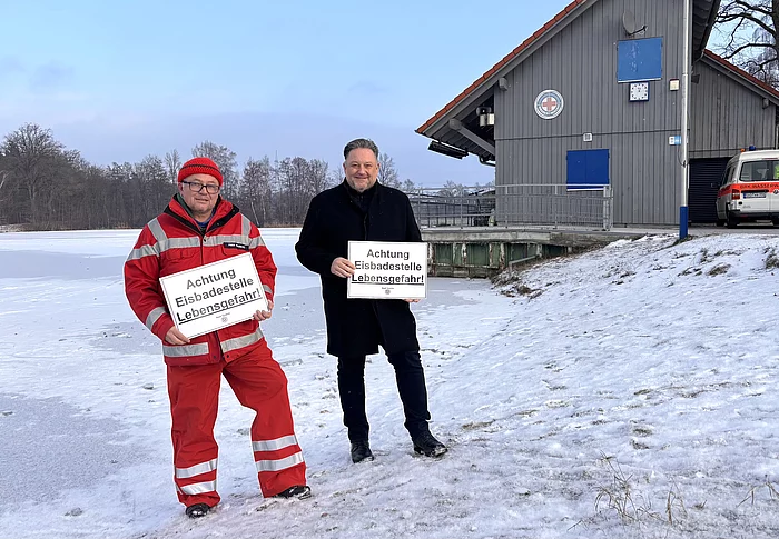Ein Vertreter der Wasserwacht und der Erste Bürgermeister warnen an einem zugefrorenen Gewässer eindringlich vor den lebensgefährlichen Risiken von Eisflächen.