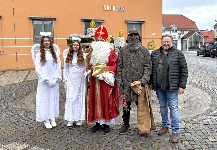 Vor dem orangefarbenen Rathaus stehen zwei als Engel verkleidete junge Personen, ein Nikolaus, eine als Knecht Ruprecht verkleidete Person sowie ein Mann in Alltagskleidung. Alle posieren für ein Gruppenfoto auf dem gepflasterten Vorplatz, wobei die festliche Kleidung auf eine Nikolausaktion hinweist.