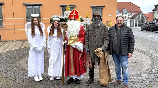 Vor dem orangefarbenen Rathaus stehen zwei als Engel verkleidete junge Personen, ein Nikolaus, eine als Knecht Ruprecht verkleidete Person sowie ein Mann in Alltagskleidung. Alle posieren für ein Gruppenfoto auf dem gepflasterten Vorplatz, wobei die festliche Kleidung auf eine Nikolausaktion hinweist.
