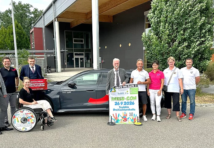 Vor der Dreifachsporthalle in Teublitz steht eine Gruppe von neun Personen neben einem dunklen Elektrofahrzeug. Eine Person sitzt im Rollstuhl. Mehrere Beteiligte halten ein Veranstaltungsplakat mit der Aufschrift „Inklu-Con 2026“ sowie einen Gutschein für einen Sportwagen. Die Gruppe lächelt in die Kamera. Das Foto entstand bei der offiziellen Vorstellung der Inklu-Con 2026, einer inklusiven Veranstaltung im Städtedreieck.