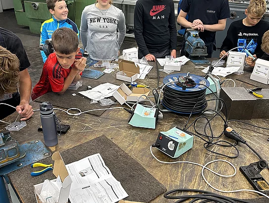 Kinder und Jugendliche arbeiten gemeinsam mit einem Betreuer an einer großen Werkbank. Darauf liegen Lötkolben, Kabel, Zangen, Bauteile und Arbeitsanleitungen.