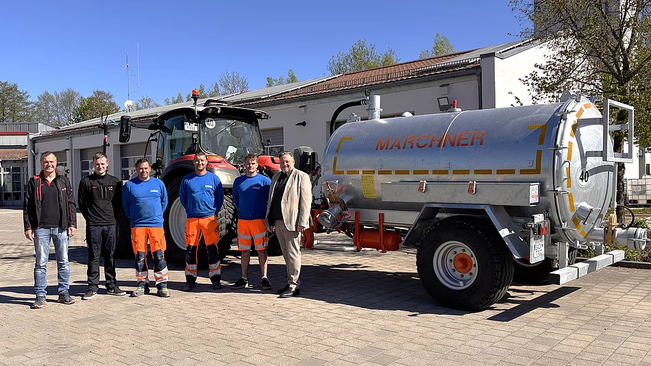 Mehrere Männer stehen bei Sonnenschein vor einem Bauhof und präsentieren gemeinsam einen Traktor mit Gießanhänger.