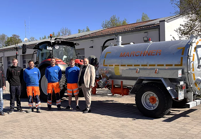 Mehrere Männer stehen bei Sonnenschein vor einem Bauhof und präsentieren gemeinsam einen Traktor mit Gießanhänger.