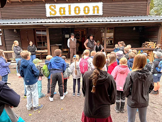 Eine größere Gruppe von Kindern und erwachsenen Begleitpersonen steht vor einem rustikalen Holzgebäude im Westernstil. Über dem Eingang befindet sich ein großes Schild mit der Aufschrift „Saloon“. Vor dem Gebäude begrüßen mehrere als Cowboys gekleidete Männer die Gruppe.