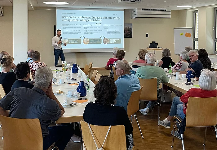 Ein Mann steht während seines Vortrags vor einer Präsentationsfläche, während zahlreiche Besucherinnen und Besucher an Tischen im Mehrgenerationenhaus sitzen und seinem Vortrag folgen.
